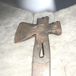 May include: A vintage, rusty metal combination tool, featuring an axe head on one side and a pry bar on the other. The tool has a cross-shaped design with a handle. The tool is resting on a textured, gray surface.