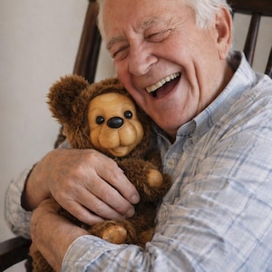 May include: A brown teddy bear with a smiling face is held by a person wearing a blue and white plaid shirt. The teddy bear has a light brown face, black eyes, and a black nose. The person is smiling broadly.
