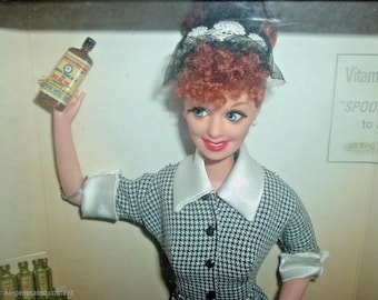 May include: A doll dressed in a black and white houndstooth print dress, holding a small brown bottle with a yellow label. The doll has red curly hair and is wearing a black hat with white lace. The doll is smiling.