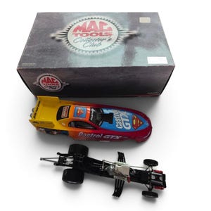 May include: A die-cast model dragster featuring a yellow, orange, and red gradient body with Castrol GTX and Superman logos, alongside a black metal chassis and a rectangular Mac Tools Collector's Club box with a dark, textured design.