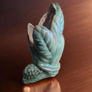May include: A ceramic vase in the shape of a stylized leaf, with a light teal glaze. The vase has a textured surface with detailed veining and a rounded base. The interior is white, contrasting with the exterior. A decorative piece for home decor.