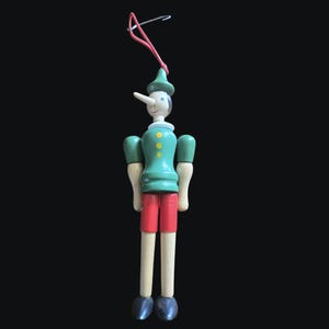 Könnte beinhalten: Eine Holzpuppe von Pinocchio mit langer Nase, die einen grünen Hut und eine Jacke, rote Shorts und schwarze Schuhe trägt. Die Puppe hat ein weißes Gesicht und wird von einer roten Schnur und einem Draht gehalten.
