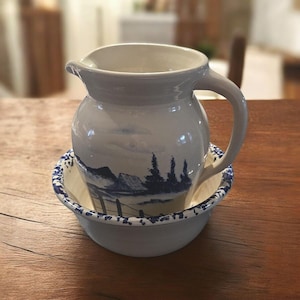 以下が含まれることがあります： 取っ手と注ぎ口が付いた白いセラミックピッチャーが、お揃いのボウルに入っています。ピッチャーとボウルには、山と木々が描かれた青い風景のデザインが施されています。ボウルには青い模様の縁があります。