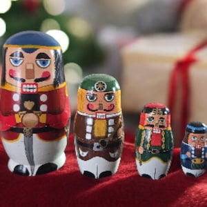May include: Four wooden nutcracker dolls of varying sizes, painted with detailed faces and costumes. The largest doll wears a red coat and blue hat. The dolls are arranged on a red surface, with a blurred background of Christmas lights and a gift box.