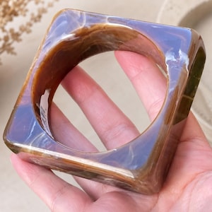 Puede incluir: Un brazalete cuadrado con apariencia marmolada. El brazalete presenta una mezcla de tonos marrón, ámbar y blanco, con un acabado brillante. El diseño es moderno y geométrico, adecuado para el uso diario.
