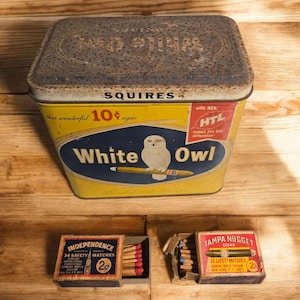 May include: An antique White Owl cigar tin with a yellow and blue design, featuring an owl illustration. Two matchboxes, one labeled "Independence" and the other "Tampa Nugget," are also visible. The items are on a wooden surface.