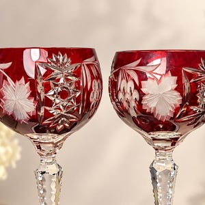 May include: Two ruby red crystal wine glasses featuring intricate hand-cut floral and geometric star patterns. The bowls are deep red with clear glass accents, supported by faceted, clear crystal stems that taper toward the base.