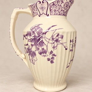 May include: A cream-colored ceramic pitcher featuring a vertical ribbed body and a purple transferware pattern. The design includes detailed botanical illustrations of leaves and flowers, with a decorative floral border at the rim.