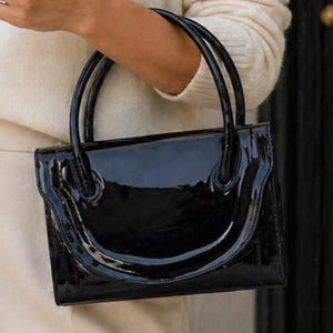 Puede incluir: Un bolso negro brillante con dos asas curvas. El bolso rectangular tiene un acabado brillante, similar al charol. El bolso lo sostiene una persona que lleva un jersey y pantalones color crema.