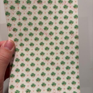 Może przedstawiać: Prostokątny kawałek papieru z powtarzającym się wzorem zielonych koniczynek i małych r&oacute;żowych kropek na kremowym tle. Papier jest trzymany przez dłoń.
