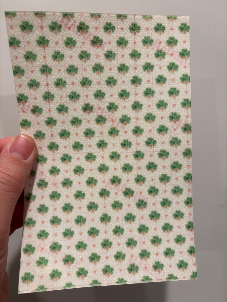 Może przedstawiać: Prostokątny kawałek papieru z powtarzającym się wzorem zielonych koniczynek i małych r&oacute;żowych kropek na kremowym tle. Papier jest trzymany przez dłoń.