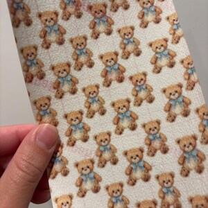 May include: A sheet of material with a repeating pattern of brown teddy bears wearing blue bows. The bears are set against a light background. The material is being held by a person.