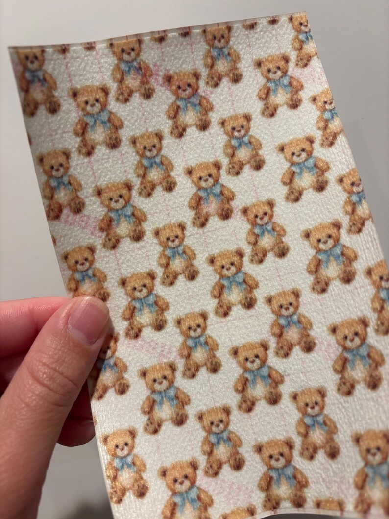 May include: A sheet of material with a repeating pattern of brown teddy bears wearing blue bows. The bears are set against a light background. The material is being held by a person.