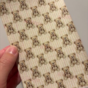 May include: A sheet of paper with a repeating pattern of small teddy bears. The bears are light brown with darker brown accents and bows. The background has a light pink and white striped design.