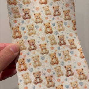 May include: A sheet of material with a repeating pattern of teddy bears in various shades of brown. The bears are interspersed with small blue and pink hearts on a white background. The material is held by a hand.