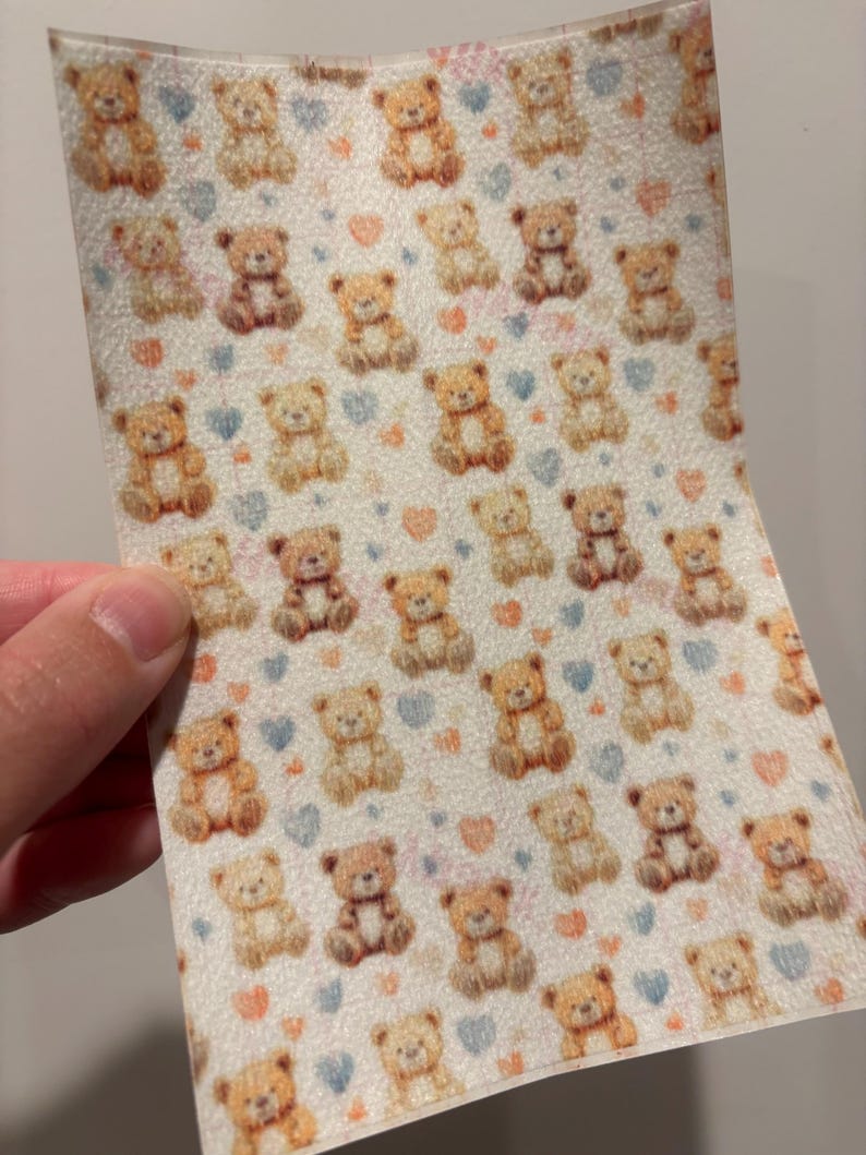 May include: A sheet of material with a repeating pattern of teddy bears in various shades of brown. The bears are interspersed with small blue and pink hearts on a white background. The material is held by a hand.