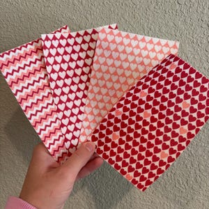 May include: A hand holds a fan of patterned paper sheets. The sheets display various designs, including red and white hearts, a pink gradient of hearts, and a red and pink zigzag pattern. The color palette is primarily red and pink.