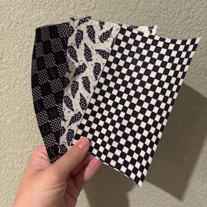 May include: Four fabric swatches with different patterns are displayed. The patterns include a black and white checkerboard, a black and white pattern with lightning bolts, and a black and white pattern with smiley faces and stars. The swatches are held in a hand.
