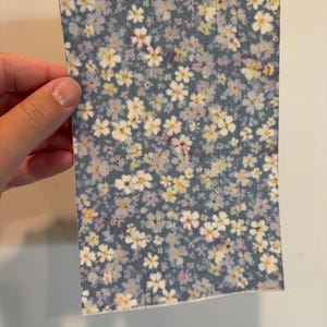 May include: A rectangular piece of fabric with a floral pattern. The fabric has a dark blue background with small white, yellow, and pink flowers scattered across the surface. The fabric is held by a hand.