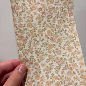 May include: A rectangular piece of paper with a floral pattern. The design features small, peach-colored flowers and brown leaves on a cream-colored background. The paper is held by a hand.
