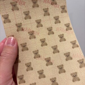 May include: A sheet of beige paper with a repeating pattern of small brown teddy bears. The paper has a grid pattern and faint pink text. The paper is being held up by a hand.