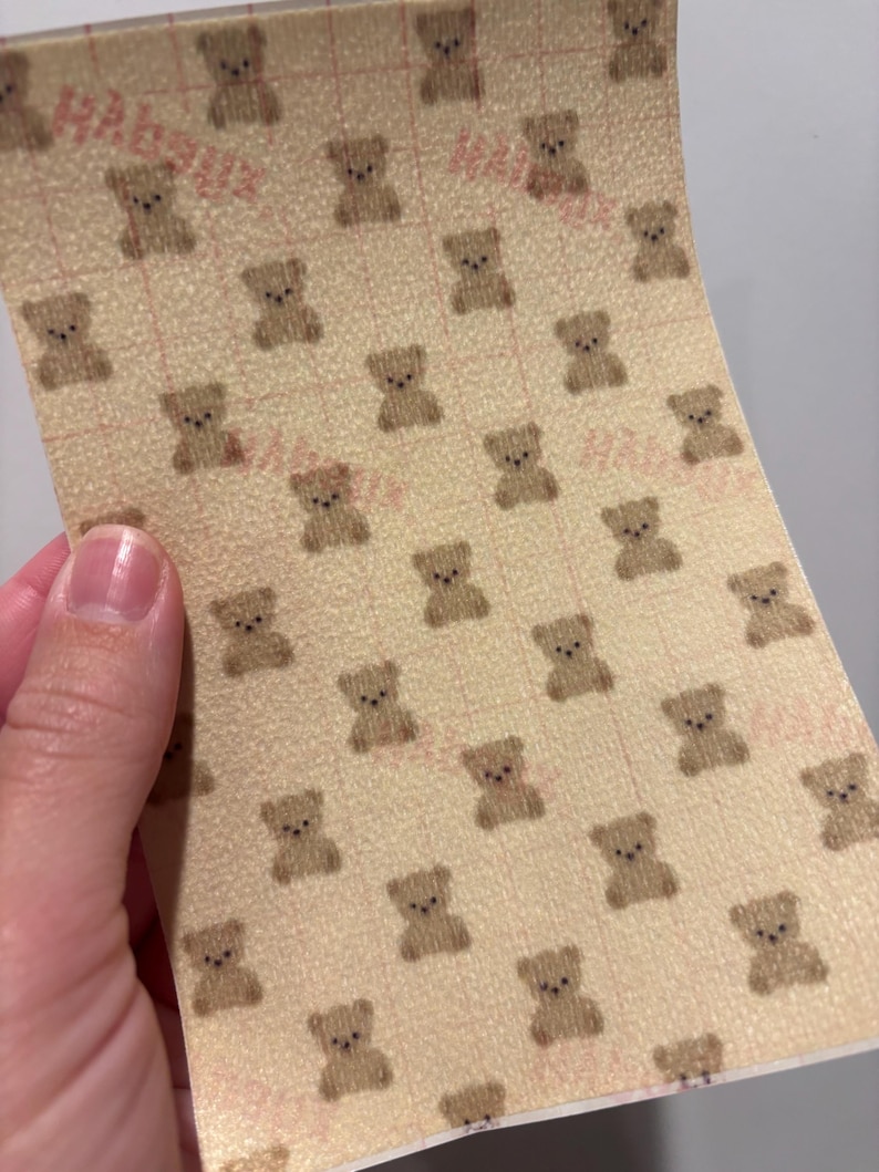 May include: A sheet of beige paper with a repeating pattern of small brown teddy bears. The paper has a grid pattern and faint pink text. The paper is being held up by a hand.
