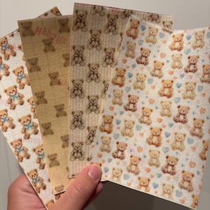 May include: A hand holds five sheets of patterned paper. Each sheet features a repeating pattern of teddy bears in various colors and designs. Some bears have blue bows. The backgrounds vary from white to yellow.