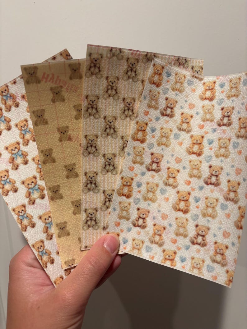 May include: A hand holds five sheets of patterned paper. Each sheet features a repeating pattern of teddy bears in various colors and designs. Some bears have blue bows. The backgrounds vary from white to yellow.