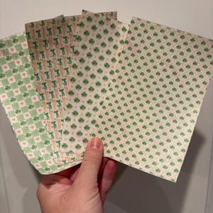 Może przedstawiać: Dłoń trzymająca kilka arkuszy papieru w wzory. Wzory obejmują zielone koniczyny, serca i powtarzający się wz&oacute;r osoby. Papier ma kremowe tło z r&oacute;żowymi i zielonymi akcentami.