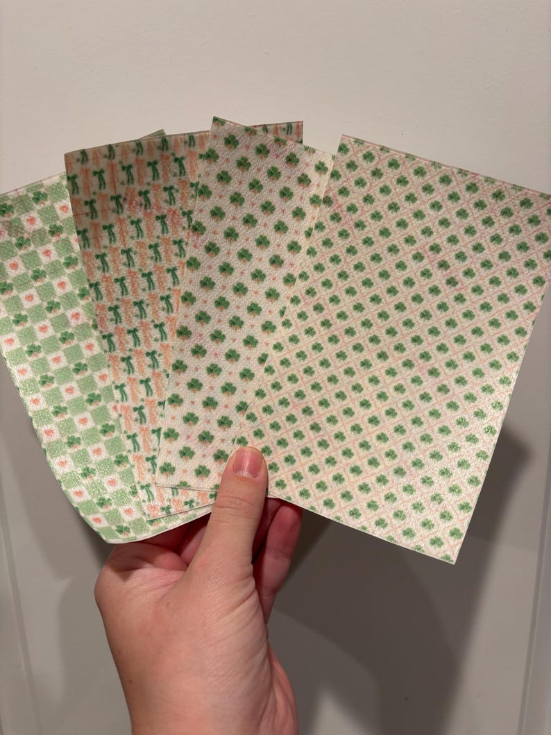 Może przedstawiać: Dłoń trzymająca kilka arkuszy papieru w wzory. Wzory obejmują zielone koniczyny, serca i powtarzający się wz&oacute;r osoby. Papier ma kremowe tło z r&oacute;żowymi i zielonymi akcentami.
