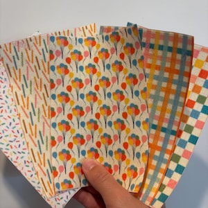 May include: A hand holds a fan of colorful patterned paper sheets. Designs include colorful sprinkles, balloon clusters, and checkered patterns in orange, blue, and pink. The sheets have a cream-colored background.