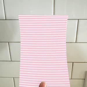 May include: A pink and white striped paper held by a hand. The paper has a rectangular shape with curved edges. The background is a white tiled wall.
