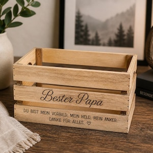Puede incluir: Una caja rectangular de madera de color marrón claro con listones abiertos. El panel frontal presenta el texto grabado "Bester Papa" en una fuente cursiva, seguido del texto "DU BIST MEIN VORBILD. MEIN HELD. MEIN ANKER. DANKE FÜR ALLES." y un corazón.