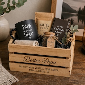May include: A wooden gift crate engraved with 'Bester Papa' and 'DU BIST MEIN VORBILD. MEIN ANKER. MEIN ZUHAUSE.' containing a black mug with 'PAPA DU BIST MEIN HELD', a coffee bag labelled 'KAFFEE F&Uuml;R DICH', a small jar with a 'F&uuml;r dich' tag, and a dark book.