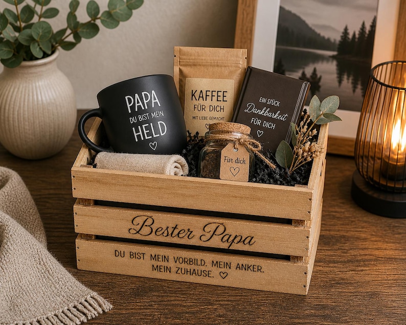May include: A wooden gift crate engraved with 'Bester Papa' and 'DU BIST MEIN VORBILD. MEIN ANKER. MEIN ZUHAUSE.' containing a black mug with 'PAPA DU BIST MEIN HELD', a coffee bag labelled 'KAFFEE F&Uuml;R DICH', a small jar with a 'F&uuml;r dich' tag, and a dark book.
