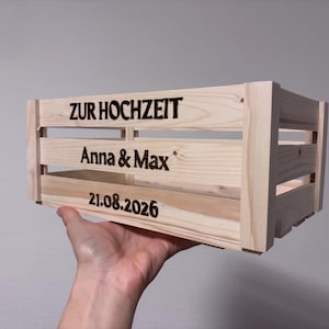 May include: A light-coloured wooden crate featuring dark, laser-engraved text on the front slats. The text reads ZUR HOCHZEIT, followed by the names Anna & Max, and the date 21.08.2026. The crate has a slatted design with visible wood grain.