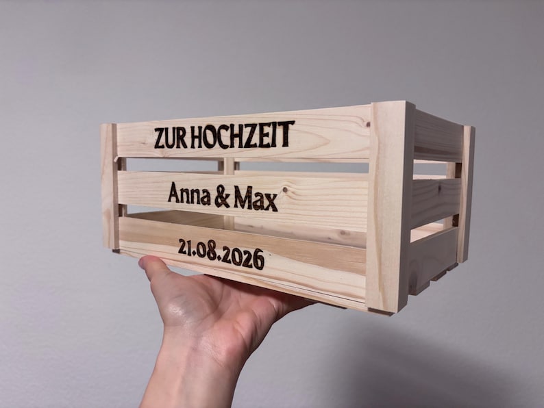 May include: A light-coloured wooden crate featuring dark, laser-engraved text on the front slats. The text reads ZUR HOCHZEIT, followed by the names Anna & Max, and the date 21.08.2026. The crate has a slatted design with visible wood grain.