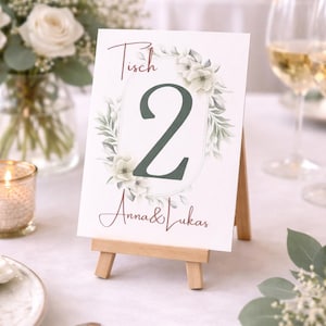Könnte beinhalten: Eine Hochzeitstisch-Nummernkarte auf einer kleinen Holzstaffelei. Die Karte zeigt die Zahl 2 in Dunkelgrün, umgeben von einem floralen Kranz. Die Namen "Anna & Lukas" sind darunter geschrieben. Das Wort "Tisch" steht über der Nummer.