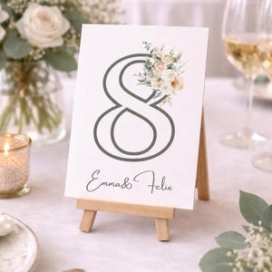 Könnte beinhalten: Eine weiße Tischnummerkarte mit einer grauen Acht und floralen Akzenten, präsentiert auf einer kleinen Holzstaffelei. Die Karte trägt die Namen "Emma & Felix" in Schreibschrift. Die Szene umfasst Blumen und Kerzen.
