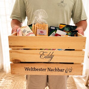 May include: A rectangular wooden crate filled with various food items, including a bag of breadsticks. The front of the crate features the name Eddy and the German text Weltbester Nachbar with two beer mug icons engraved on the bottom slat.
