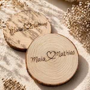 May include: Two round wooden coasters with the names "Maia" and "Mathias" engraved in a cursive font, connected by a heart symbol. The coasters are made from natural wood and have visible wood grain.