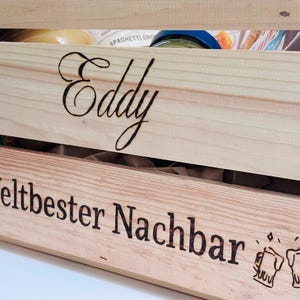 May include: A light-coloured wooden crate featuring the name Eddy engraved in a cursive script on the top slat and the text Weltbester Nachbar with two clinking beer mug icons engraved on the bottom slat, containing various food items inside.