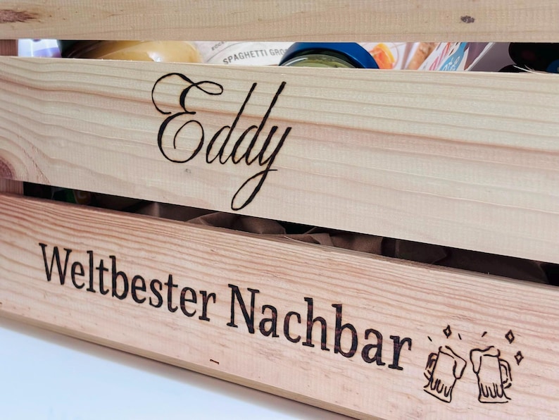 May include: A light-coloured wooden crate featuring the name Eddy engraved in a cursive script on the top slat and the text Weltbester Nachbar with two clinking beer mug icons engraved on the bottom slat, containing various food items inside.