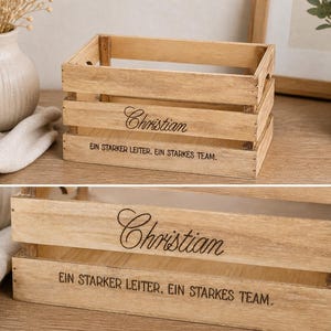 May include: A rectangular light brown wooden crate with horizontal slats. The front panel features the name Christian in a cursive script font, followed by the text EIN STARKER LEITER. EIN STARKES TEAM. printed in a simple sans-serif font.