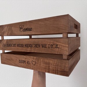 May include: A rectangular brown wooden crate with horizontal slats and cutout handles. The side features laser-engraved text reading Carina, ES BRAUCHT MEHR MENSCHEN WIE DICH., and HAPPY 40. followed by a heart symbol, showcasing a natural wood grain finish.