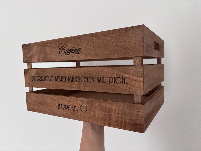 May include: A rectangular brown wooden crate with horizontal slats and cutout handles. The side features laser-engraved text reading Carina, ES BRAUCHT MEHR MENSCHEN WIE DICH., and HAPPY 40. followed by a heart symbol, showcasing a natural wood grain finish.