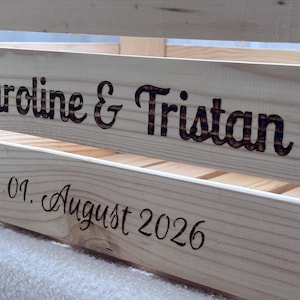 May include: A light-coloured wooden crate featuring dark, burned-in lettering. The top slat displays the names Caroline & Tristan in a bold font, while the lower slat shows the date 01. August 2026 in a cursive script style, engraved into the wood.