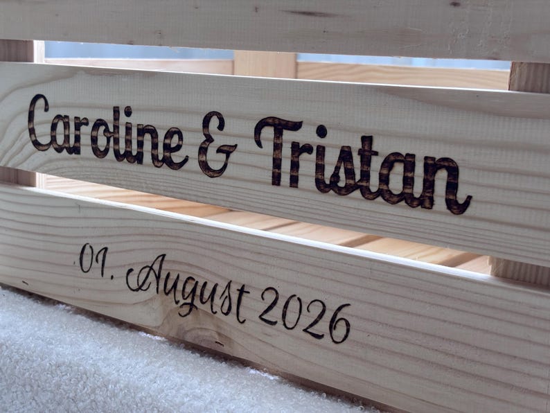 May include: A light-coloured wooden crate featuring dark, burned-in lettering. The top slat displays the names Caroline & Tristan in a bold font, while the lower slat shows the date 01. August 2026 in a cursive script style, engraved into the wood.