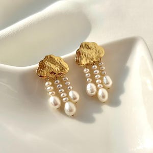 Puede incluir: Un par de pendientes dorados con forma de nube con perlas colgantes en forma de lágrima. Los pendientes se exhiben sobre una superficie blanca, mostrando su elegante diseño y delicados detalles. Las perlas son blancas y brillantes.