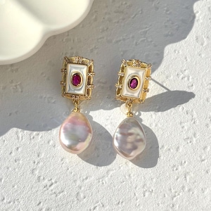 May include: A pair of gold-toned earrings with a square top featuring a white center and a small red stone. Each earring has a teardrop-shaped, iridescent pearl pendant. The earrings are displayed on a textured white surface.